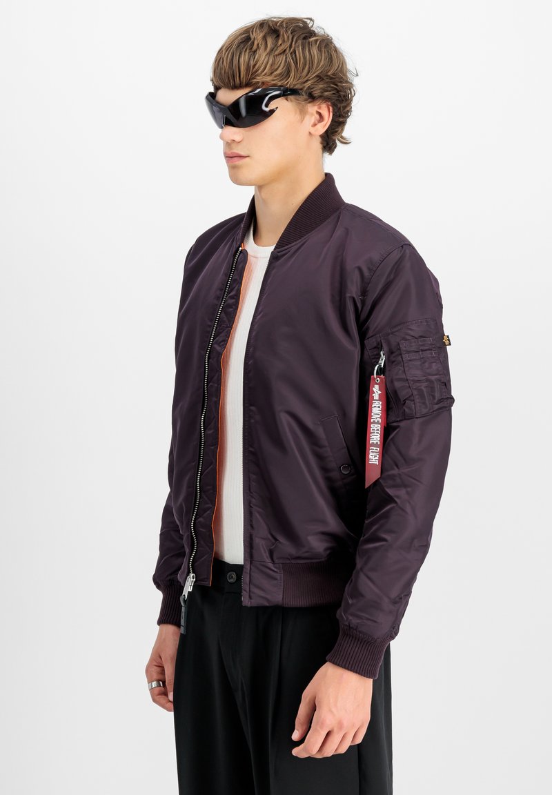 Chaqueta bomber burdeos con puños acanalados, cierre de cremallera, forro naranja vertical y un bolsillo de parche en la manga. Lleva puesta unas gafas de sol negras.