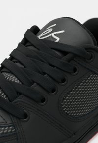 Baskets noires avec de gros lacets noirs, panneau latéral texturé et logo blanc brodé sur la languette rembourrée.