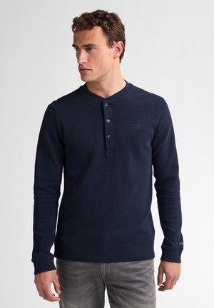 Young man wearing a dark blue long-sleeve henley shirt with three buttons, paired with light gray jeans, looking to the left.