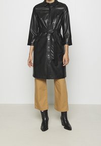 Czarna sukienka-koszula z imitacji skóry z kołnierzykiem, zapinana na guziki, z bocznymi kieszeniami i dopasowanym paskiem, zestawiona z beżowymi spodniami o szerokich nogawkach i czarnymi butami.