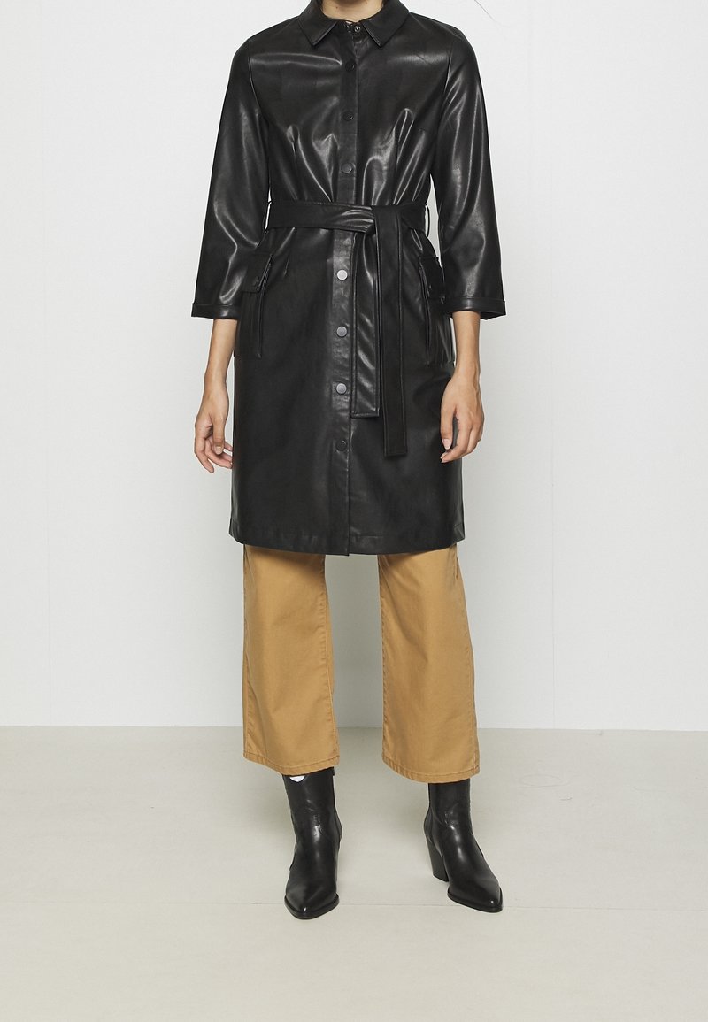 Czarna sukienka-koszula z imitacji skóry z kołnierzykiem, zapinana na guziki, z bocznymi kieszeniami i dopasowanym paskiem, zestawiona z beżowymi spodniami o szerokich nogawkach i czarnymi butami.