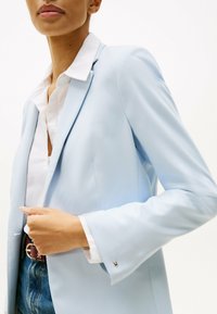 Blazer su misura blu chiaro con chiusura a un bottone, rever a punta e una texture liscia, abbinato a una camicia bianca con colletto.
