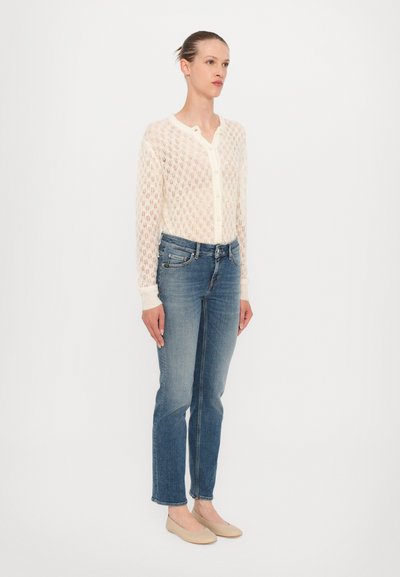 Leichter, gestrickter cremefarbener Cardigan mit einem Rautenmuster, Knopfverschluss und gerippten Bündchen, kombiniert mit blauen geraden Jeans und beigen Ballerinas.