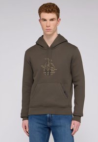 Ciemnozielona bluza z kapturem o teksturowanym materiale, złote logo na klatce piersiowej, kieszeń kangurza i ściągacze u rękawów. Zestawiona ze średnio niebieskimi dżinsami.