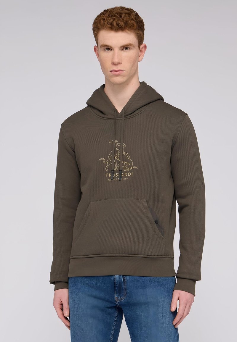 Ciemnozielona bluza z kapturem o teksturowanym materiale, złote logo na klatce piersiowej, kieszeń kangurza i ściągacze u rękawów. Zestawiona ze średnio niebieskimi dżinsami.