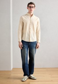 Light beige button-up shirt with a subtle texture, paired with dark blue jeans and green-accented sneakers. Simple, tailored fit.