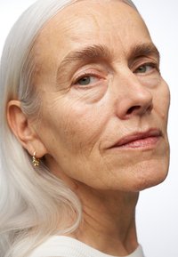Gros plan sur une femme avec de longs cheveux argentés et raides, une peau claire et des boucles d'oreilles en or. Son visage montre de fines rides et une expression neutre.