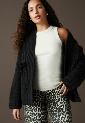 Schwarze Strickjacke mit strukturierten Mustern über einem hellgrauen, gerippten Tanktop, kombiniert mit beigen, leopardenmusternden Hosen.