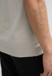 Hellgraues, gestricktes T-Shirt mit Rundhalsausschnitt, kurzen Ärmeln und geripptem Saum, kombiniert mit schwarzen Hosen. Keine auffälligen Muster festgestellt.