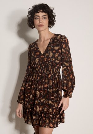 Femme aux cheveux courts et bouclés portant une robe longue à manches longues de couleur foncée avec un motif cachemire multicolore, debout devant un fond uni.