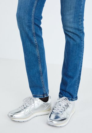 Silver metallic sneakers with a smooth texture, round toe, and white rubber sole, paired with fitted blue jeans with faint stitching.