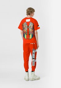 Camiseta gráfica roja con un diseño de alas colorido, combinada con pantalones rojos a juego que presentan detalles desgastados y una etiqueta. Botas gruesas blancas.