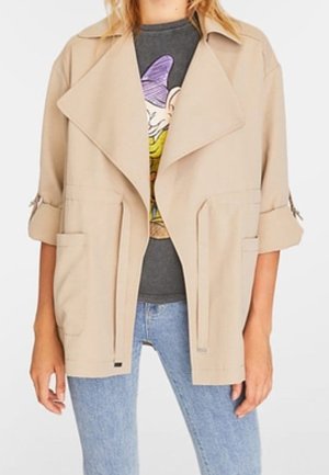 Persona che indossa una giacca beige aperta sul davanti con maniche arrotolate su una T-shirt grigia con grafica e jeans azzurri.