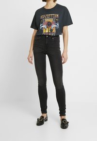 Persona con una camiseta gráfica negra de Led Zeppelin, jeans ajustados de cintura alta negros y zapatos slip-on negros con hebillas doradas.