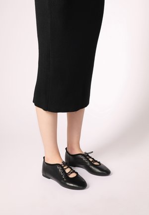 Black leather lace-up shoes with an open upper design, featuring eyelets and a flat sole, paired with a fitted black ribbed dress.
