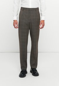 Mann in braunen karierten Anzughosen, weißem Hemd mit Knöpfen und schwarzen Loafers, der vor einem schlichten weißen Hintergrund steht.