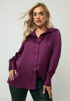 Blonde vrouw met golvend haar, gekleed in een paarse satijnen blouse met lange mouwen en een zwarte broek, staand tegen een effen lichte achtergrond.