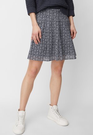 Persoon met een A-lijn rok in marineblauw met patroon, een marineblauwe gebreide trui en witte hoge sneakers, staand met de armen langs het lichaam.