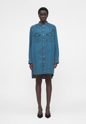 Femme portant une robe-chemise en denim bleu à manches longues boutonnée avec poches à rabat, debout, droite, devant un fond clair uni.