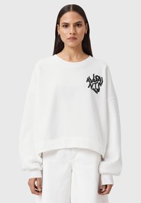 Una donna con lunghi capelli scuri indossa una felpa oversize bianca con il logo nero "AllSaints London" sul petto e pantaloni bianchi a gamba larga.