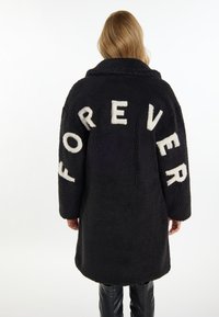 Černý kabát z umělé kožešiny s velkým bílým nápisem "FOREVER" na zadní straně, s límcem, střední délky a dlouhými rukávy.
