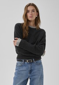 Dunkelgrauer Strickpullover mit einem hellgrauen, gerippten Rundhalsausschnitt und Bündchen. Kombiniert mit mittelblauen Jeans und einem schwarzen Gürtel mit silberner Schnalle.