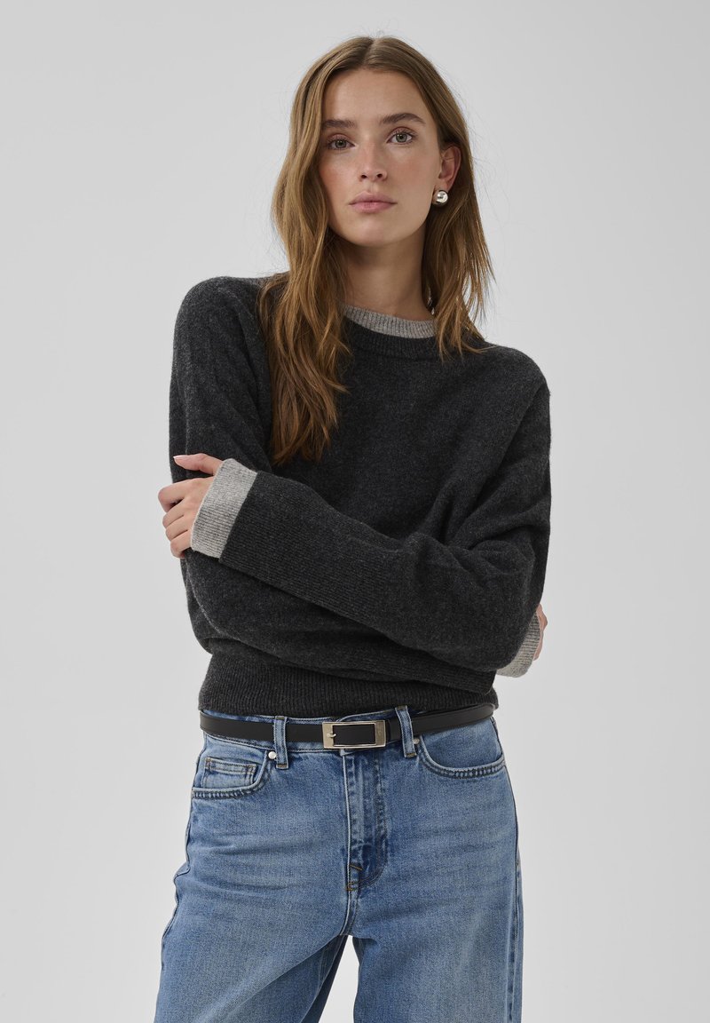 Dunkelgrauer Strickpullover mit einem hellgrauen, gerippten Rundhalsausschnitt und Bündchen. Kombiniert mit mittelblauen Jeans und einem schwarzen Gürtel mit silberner Schnalle.