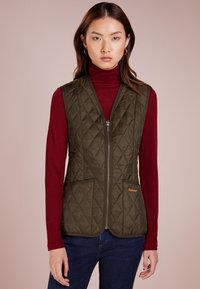 Gepolsterte olivgrüne Weste mit einem Reißverschluss, ausgestattet mit zwei Fronttaschen und braunen Kanten, getragen über einem roten Rollkragenpullover und blauen Jeans.
