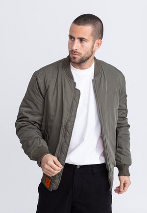 Man die de rits vastmaakt van een olijfgroene bomberjas, draagt een wit overhemd en een zwarte broek, kijkt opzij tegen een effen achtergrond.