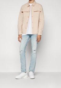 Chaqueta de camisa de manga larga en un beige claro con dos bolsillos en el pecho, combinada con jeans desgastados de un azul claro y zapatillas blancas.