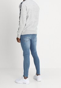 Person som bär en ljusgrå sweatshirt med svart logotape på ärmarna, ljusblå slim-fit jeans och vita sneakers, stående mot en vit bakgrund.