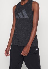 Canotta atletica nera senza maniche con una texture a macchie, caratterizzata da un grande logo Adidas grigio sul davanti. Abbinata a pantaloni neri.