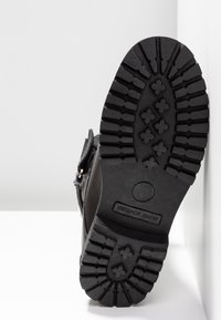 Semelle de botte noire avec un motif de bande de roulement robuste ; comporte le logo "Versace Jeans" en relief et une surface plane pour l'adhérence.