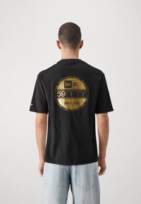 T-shirt in cotone nero con un grande logo circolare dorato sul retro, con il testo "59FIFTY" e "SINCE 1920". Maniche corte, vestibilità rilassata.