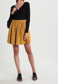 Top negro de manga larga cruzado; falda amarilla con pliegues; zuecos negros destalonados. Textura suave; cuerpo ajustado; falda acampanada; cinturilla fruncida.