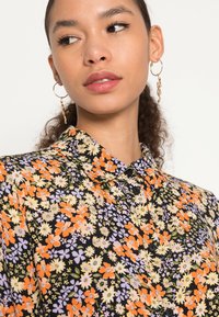 Blusa floral com flores multicoloridas em laranja, amarelo e roxo sobre um fundo preto; apresenta um decote com colarinho e botões pretos.