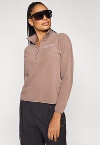 Sudadera marrón de forro polar con cremallera de cuarto, mangas largas y un ajuste cómodo; presenta un logo sutil en el pecho. Material texturizado y suave.