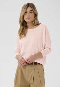 Pull en maille rose clair avec une coupe décontractée, un large col bateau et des poignets de manches côtelés, associé à un pantalon taille haute kaki.