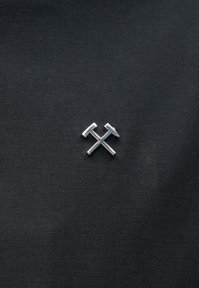 Μικρό ασημένιο καρφίτσα σε σχήμα δύο σταυρωτών σφυριών πάνω σε μαύρο υφασμάτινο φόντο με υφή.