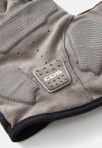 Manusă gri cu un aspect texturat din suede și accente de cauciuc, cu perforații și un patch cu logo. Designul include segmente flexibile pentru degete.