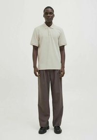 Polo beige de manga corta, textura suave y cuello discreto. Combinado con pantalones sueltos marrón oscuro y calzado negro.