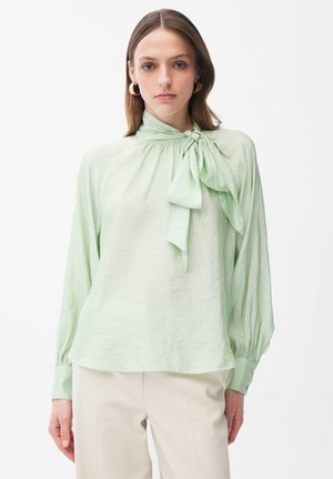 Femme portant un chemisier à manches longues vert clair avec un grand nœud au col et un pantalon crème, debout devant un fond blanc uni.