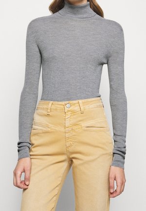 Femme portant un pull col roulé gris côtelé et ajusté, rentré dans un pantalon beige taille haute avec poches avant et passants pour ceinture.