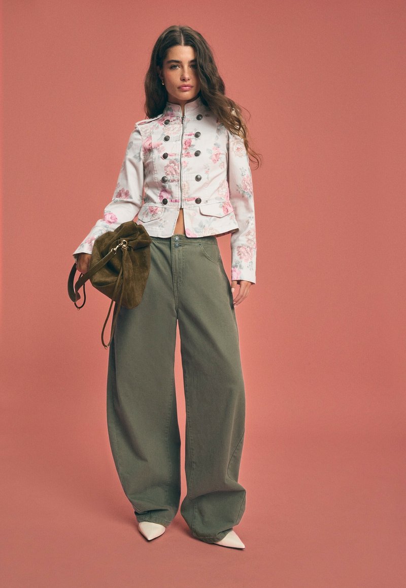 Jeune femme en veste courte fleurie, pantalon large kaki, chaussures blanches à bouts pointus, tenant un sac en daim marron, debout devant un fond rougeâtre atténué.
