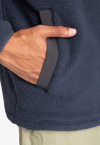 Giacca in pile blu navy con una mano in una tasca laterale, bordi grigi a contrasto lungo l'apertura della tasca e superficie in tessuto testurizzato.