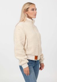 Giacca in pile beige con colletto alto, zip frontale e polsini elasticizzati. Presenta un aspetto texture e un'etichetta sulla vita.