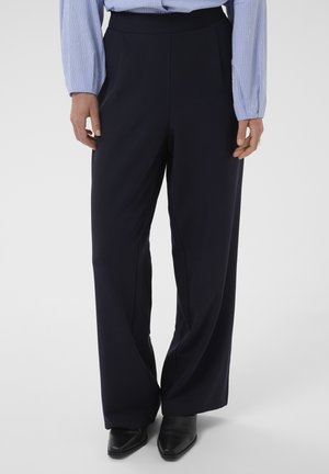 Femme portant un pantalon noir taille haute à jambes larges, une chemise à carreaux bleu clair et des chaussures noires pointues, debout devant un fond uni.