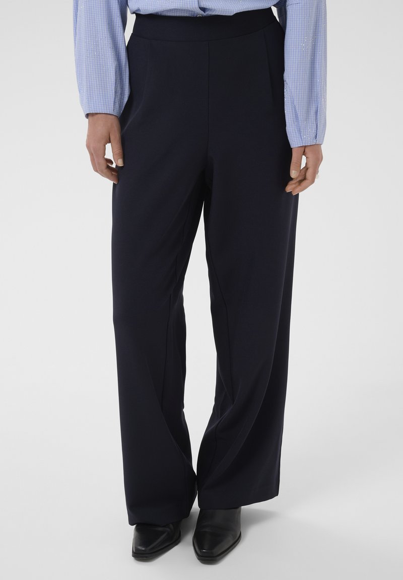 Femme portant un pantalon noir taille haute à jambes larges, une chemise à carreaux bleu clair et des chaussures noires pointues, debout devant un fond uni.