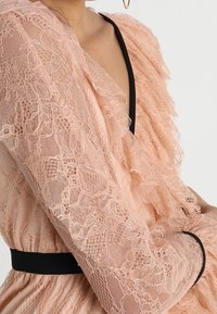 Kvinna i en persikofärgad spetsklänning med volanger, svart kant längs halsringningen och midjan, samt guldörhängen, med armarna i kors.