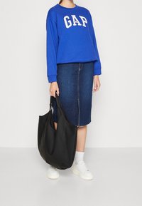 Sudadera azul con el logo "GAP", falda de lápiz de mezclilla, bolsa tote negra, zapatillas blancas y calcetines tobilleros. Conjunto simple y casual.